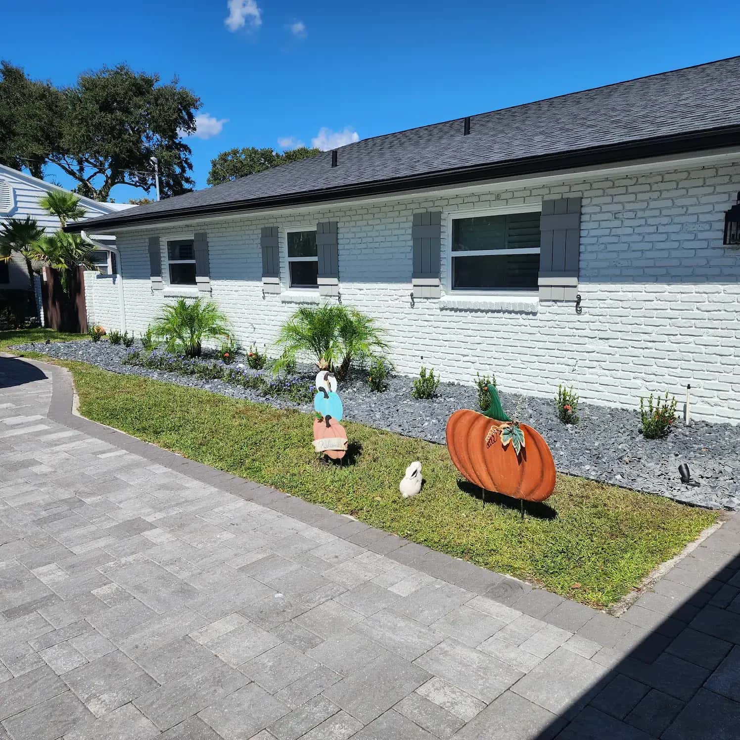 Gray paver driveway and walkway beside a white brick home with slate rock beds, palms and a strip of lawn